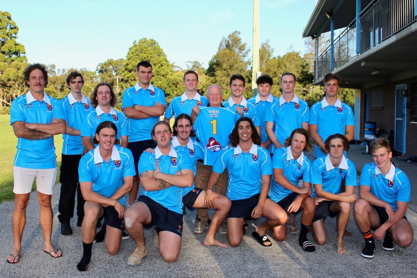 The Wakehurst Colts in sky-blue polos gathered around Bladesie outside the clubhouse as he holds up his number 1 jersey.