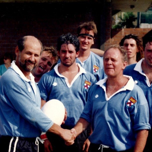 An older faded photo of Wakehurst players in sky-blue jerseys; Bladesie, on the right, shakes hands with a man holding a rugby ball.
