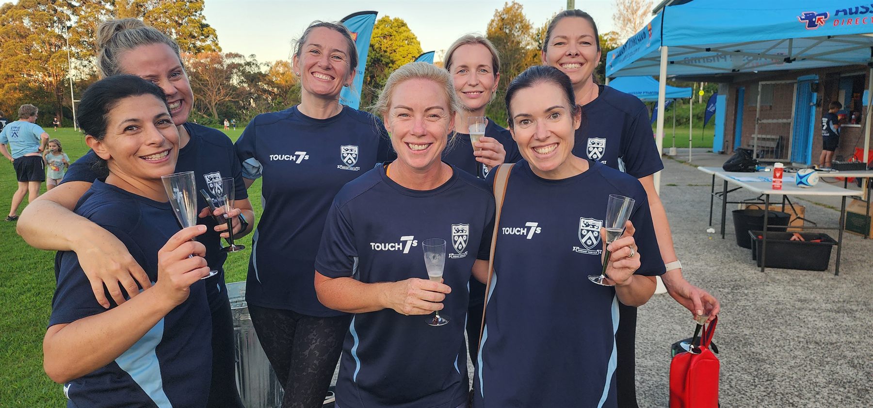 Wakehurst women in navy and light blue Touch 7s jerseys raising glasses together on the sideline at Wakehurst Rugby Park as the sun sets behind the clubhouse.