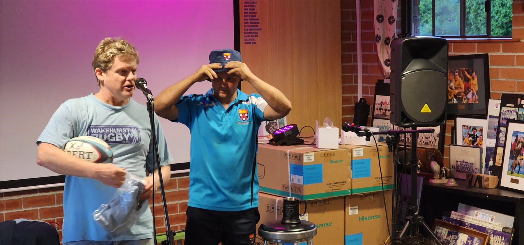 Two Wakehurst men at the microphone in the clubhouse during a presentation night, one holding a rugby ball and a folded jersey, the other pulling on a club cap, with the honour boards and framed club photographs behind them.